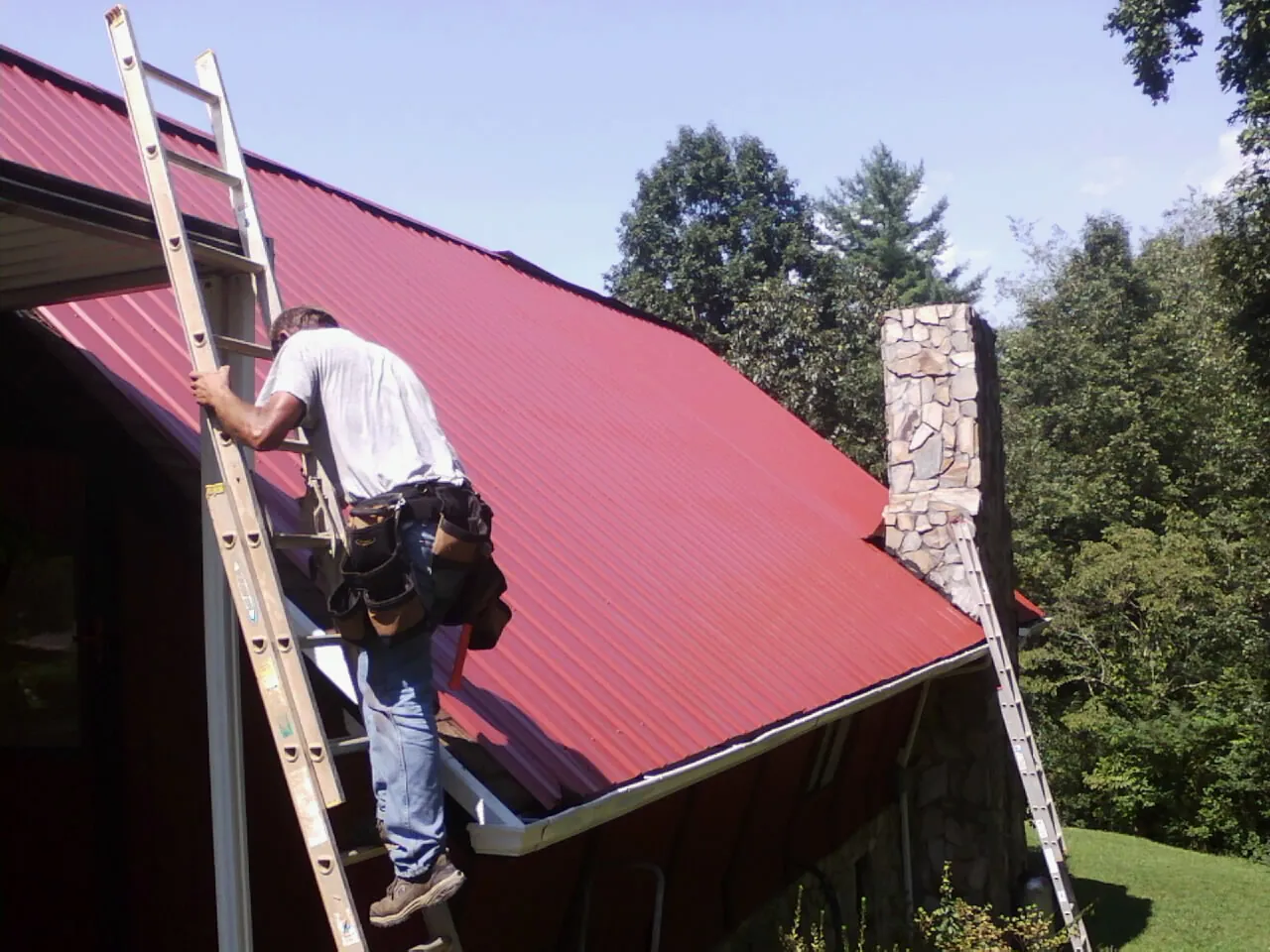 Roofer on a ladder beside a red metal roof and stone chimney — Soffit & Fascia Repair by Roofing Brothers in Rose Hill